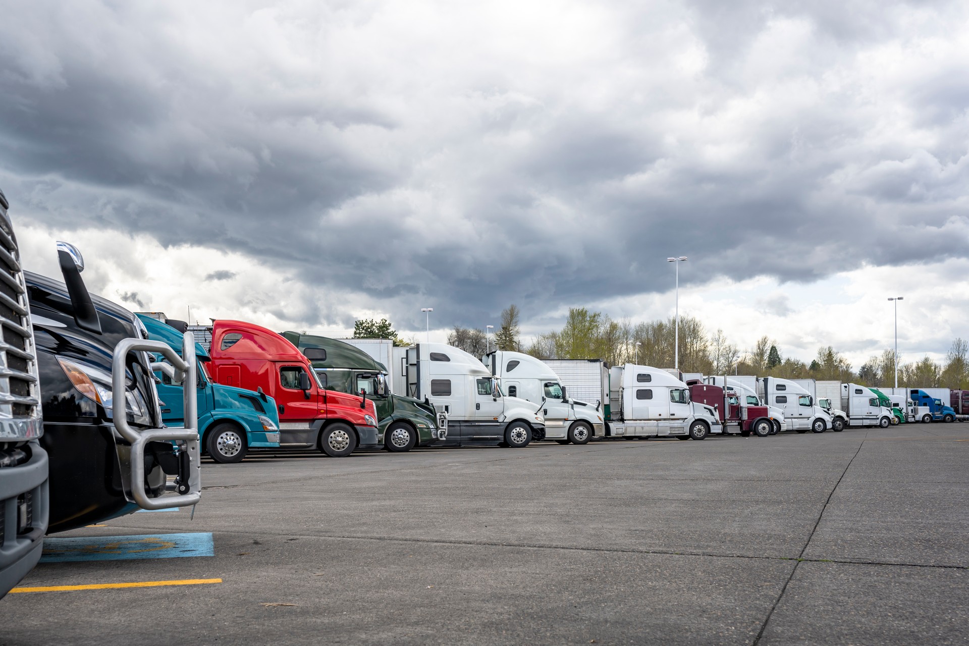 Different big rigs semi trucks with semi trailers standing in long row on truck stop parking lot with stormy sky