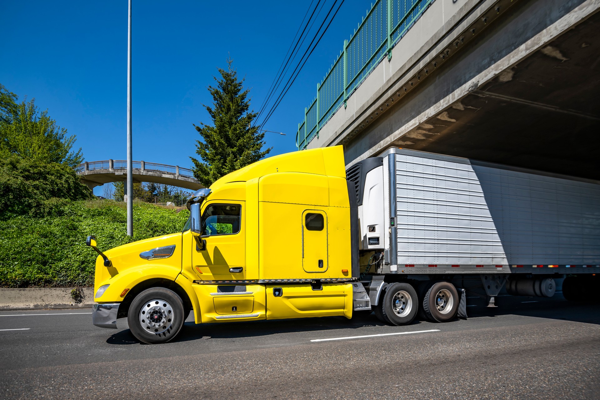 Yellow professional big rig bonnet semi truck transporting frozen cargo in refrigerator semi trailer running on the highway road under the bridge